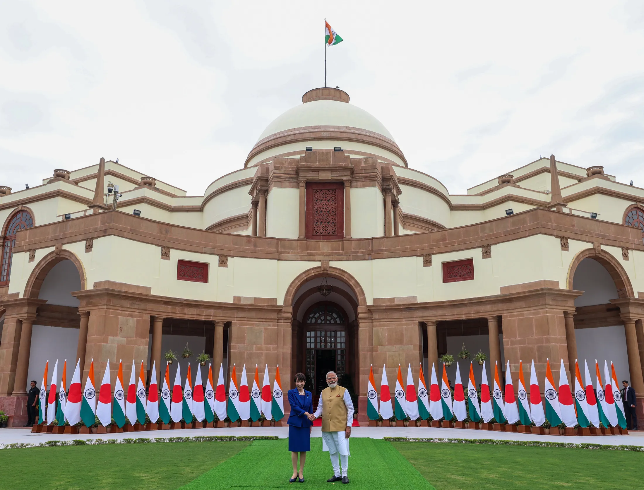 Prime Minister Narendra Modi and Japanese Prime Minister Sanae Takaichi shake hands in front of Hyderabad House, New Delhi, flanked by rows of Indian and Japanese flags.